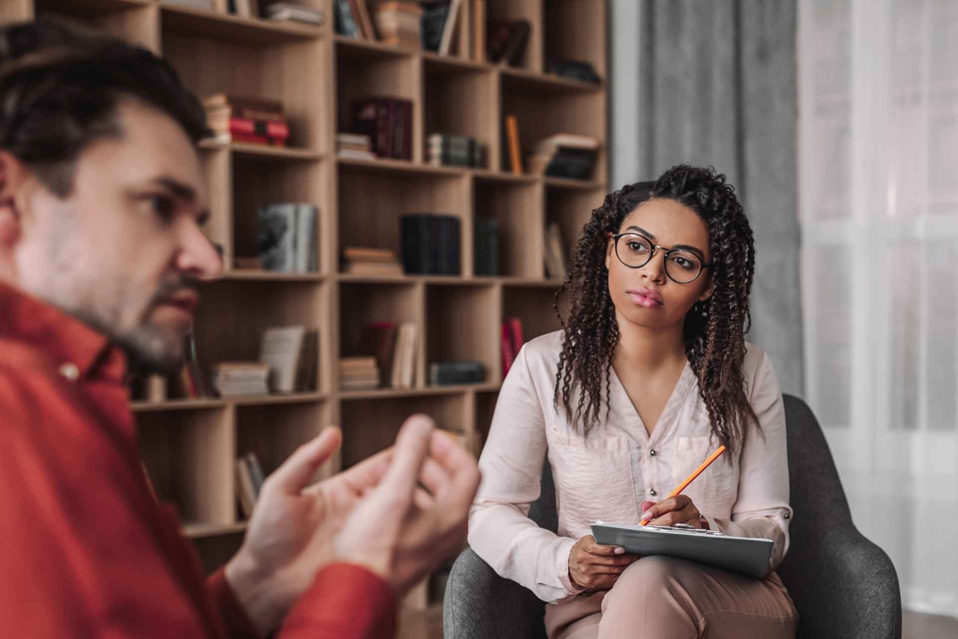 Female mental health professional holding a clipboard while listening to a man during an individual counseling session.