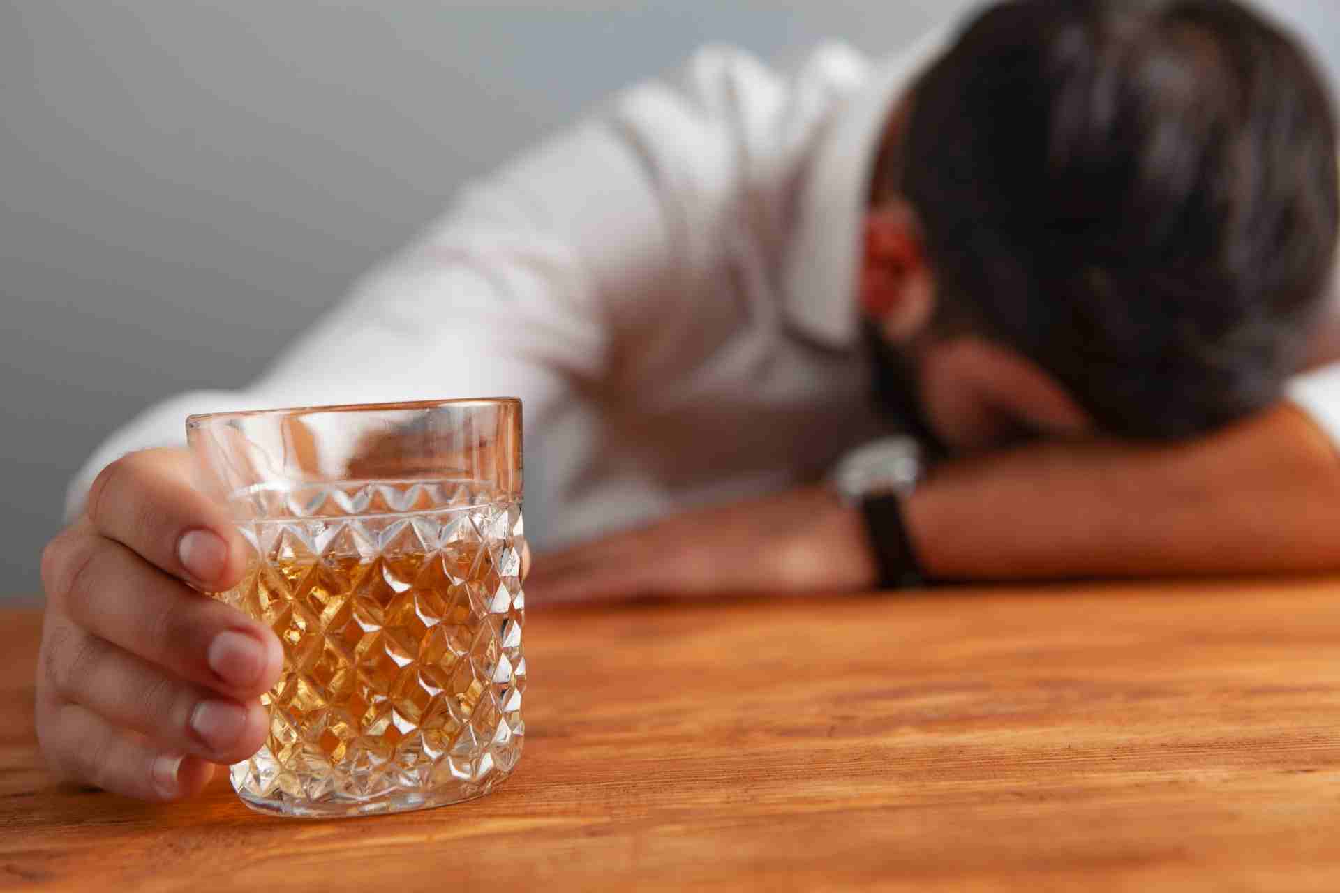 Man asleep at a table holding a glass of alcohol, illustrating a warning sign of alcohol addiction.